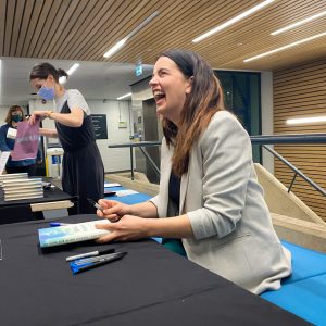 Kate Bowler is sat at a table signing copies of her book 'No Cure for Being Human' and smiling